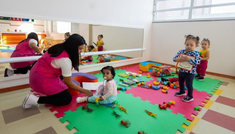 foto de bebês e uma professora em uma sala com espelho, brinquedos e tapete emborrachado colorido