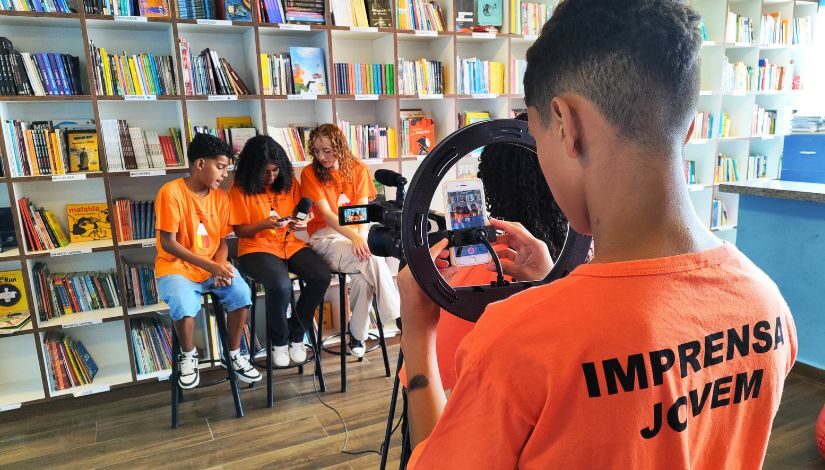 Fotografia de cinco estudantes com a camiseta laranja do Imprensa Jovem. Três estudantes estão sentados em bancos altos em frente a uma estante de livros e a menina que está no meio segura um microfone e um celular. Todos olham para o celular. Em primeiro plano, de costas para o ângulo da fotografia, um menino manuseia o tripé em que está a câmera e o celular, atrás de sua camiseta laranja está escrito em preto Imprensa Jovem. Em frente a ele, e também de costas para o ângulo da foto, há mais uma estudante.