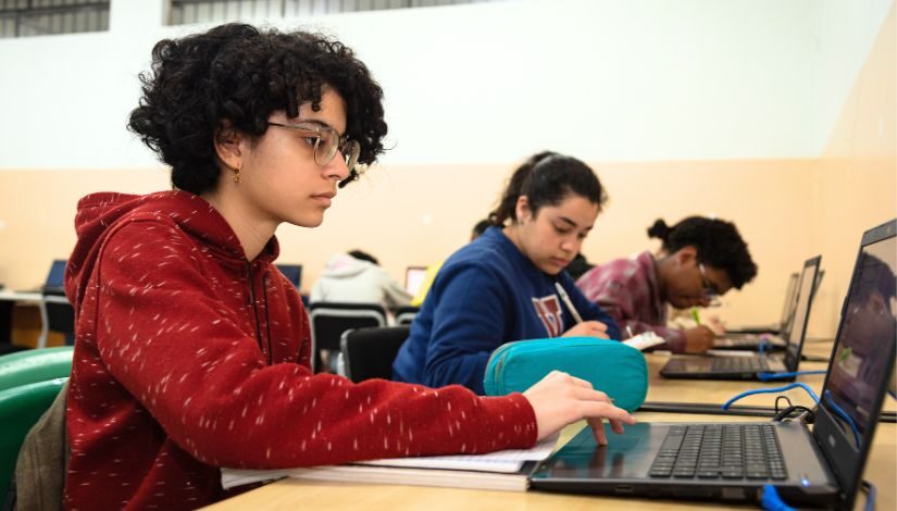 Fotografia de três estudantes do Ensino Médio da Escola Municipal de Ensino Fundamental e Médio (EMEFM) Derville Allegretti. As três estudantes estão sentadas em frente a mesas na sala de aula. A menina que está em primeiro plano olha para o computador e está com a mão no touchpad. As outras duas escrevem em seus cadernos.