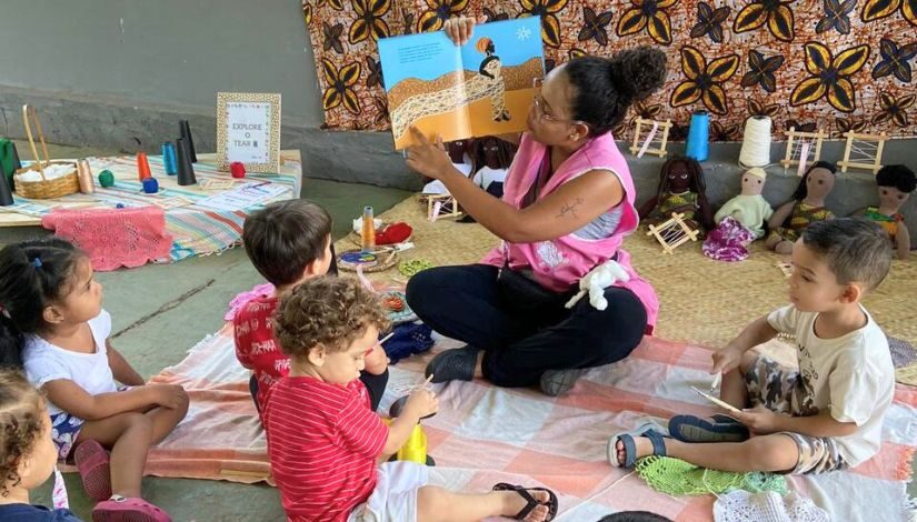 Fotografia mostra professora sentada no chão, ela segura um livro com uma mão e aponta para as figuras com a outra. Na sua frente, há crianças sentadas no chão brincando com linhas e agulhas. No fundo da foto, pode-se ver os bonecos e bonecas negros e bolivianos.