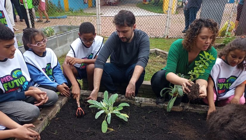 Fotografia mostra quatro estudantes e dois professores plantando em hrota. Na frente deles, há um espaço com terra, uma muda já está plantada e outra está sendo colocada.