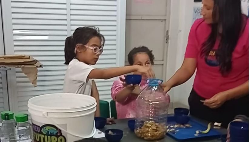 Fotografia mostra menina colocando cascas de alimentos em uma garrafa plástica que está sendo segurada por uma mulher. Atrás da garrafa há uma menina segurando um pote azul.