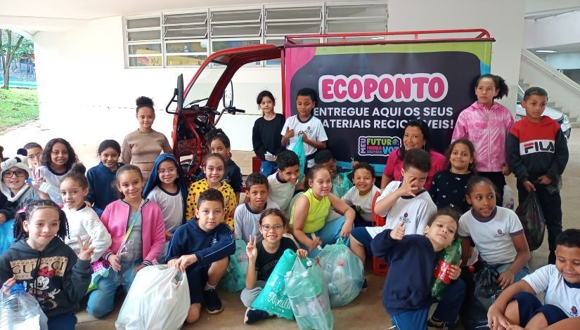 Estudantes estão posando para foto em frente a um carrinho onde se lê "Ecoponto - Entregue aqui seus materiais recicláveis".