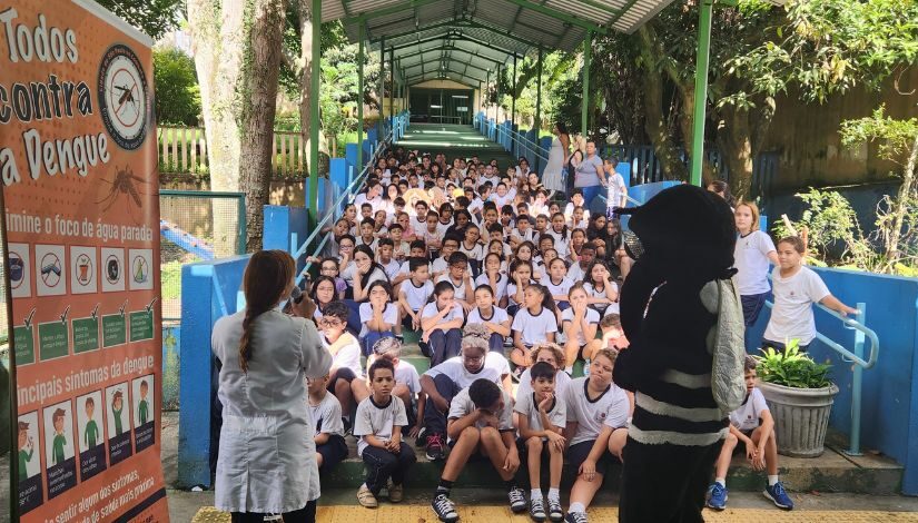 foto de estudantes sentados em uma escada, vendo explicações sobre o combate à denque. Um adulto está fantasiado de mosquito