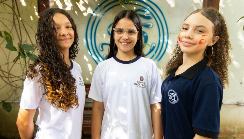 Três estidantes com uniformes da prefeitura olham para a câmera sorrindo.