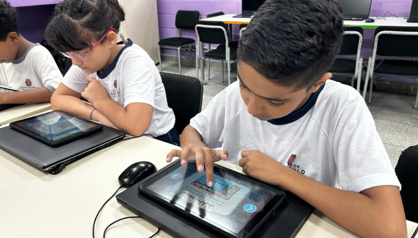 Uma criança com uniforme da prefeitura toca em um tablet em cima de uma mesa