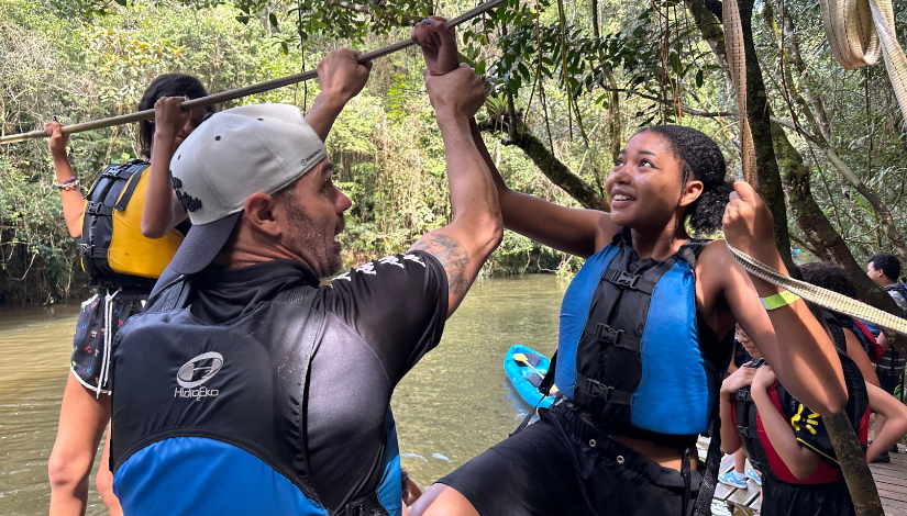Um professor segura o braço de uma estudante que sobe em uma corda. Ao fundo, uma floresta e um rio.