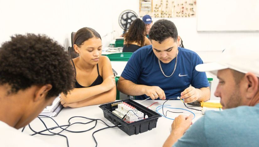 Fotografia mostra quatro pessoas adultas ao redor de uma mesa com componentes eletrônicos na aula do curso profissionalizante de auxiliar de eletricista.