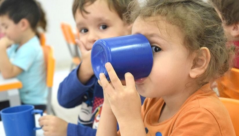 Foto de uma criança com um copo na copa bebendo algo. Atrás dela há outras crianças.