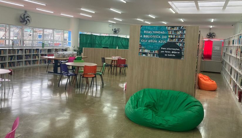 Fotografia mostra o ambiente de uma biblioteca, com estante de livros, mesas e cadeiras e puffs.