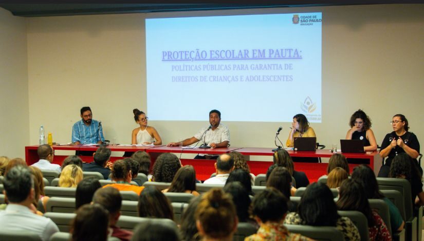 Fotografia mostra seis pessoas sentadas em uma bancada vermelha, atrás deles se lê "Proteção escolar em pauta: Políticas públicas para garantia de direitos de crianças e adolescentes". Na frente deles, há diversas pessoas sentadas.