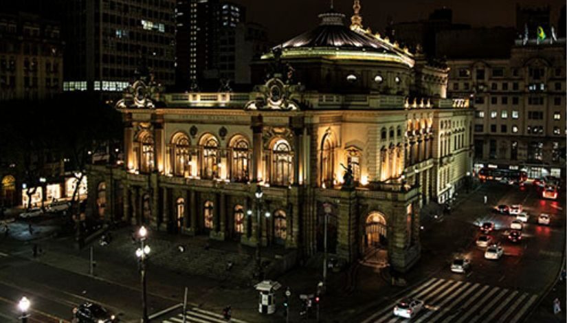 Fotografia do Theatro Municipal da parte externa toda iluminada durante a noite.