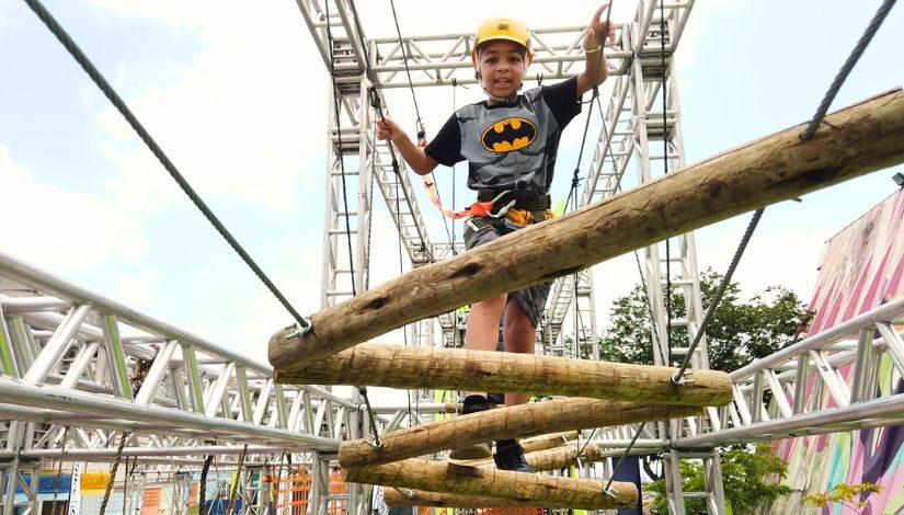 Fotografia mostra menino com capacete em um trilha com troncos de madeira suspensa.