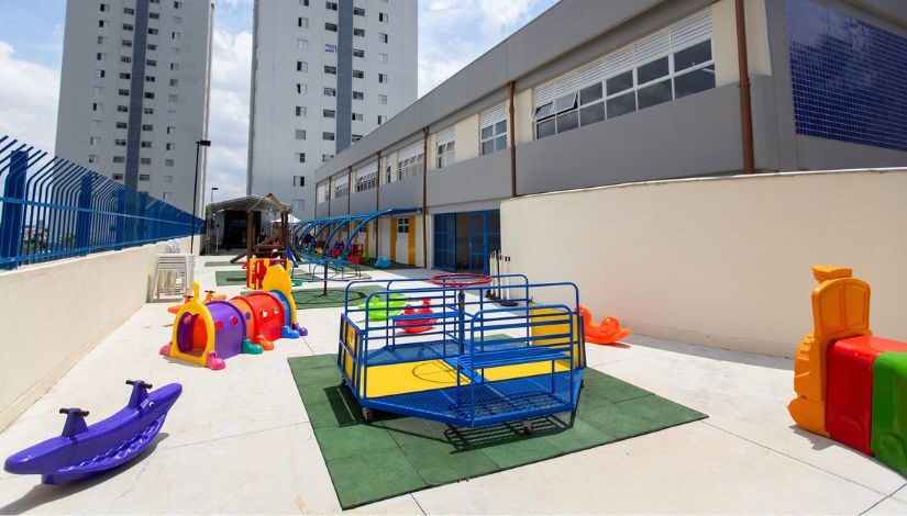 Fotografia mostra área externa de um Centro de Educação Infantil (CEI) com brinquedos do parque, como gira-gira e túnel de centopeia.