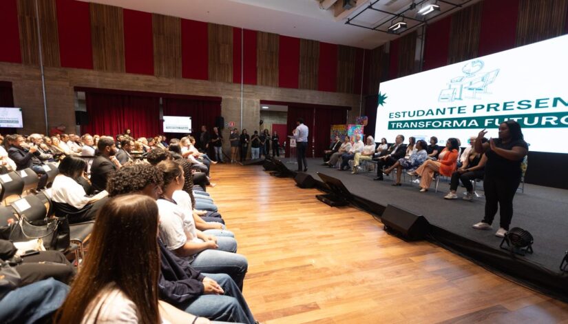 Fotografia de várias pessoas em um auditório assistindo uma apresentação da campanha "Estudante Presente Transforma Futuros"