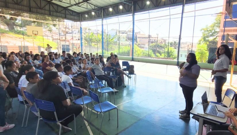 Fotografia com vários estudantes sentados em cadeiras verdes na quadra coberta da escola.