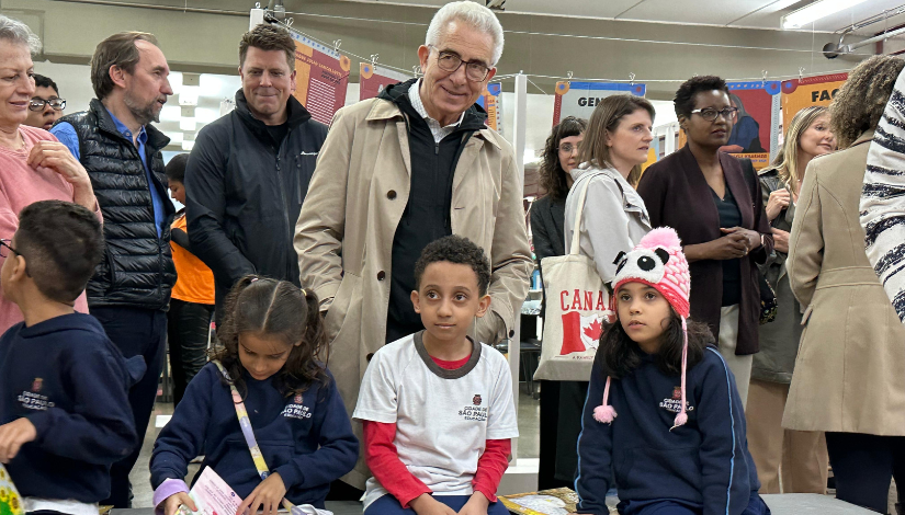 Três crianças sentadas em bancos lendo livro, ao fundo, um grupo de adultos conversa em pé