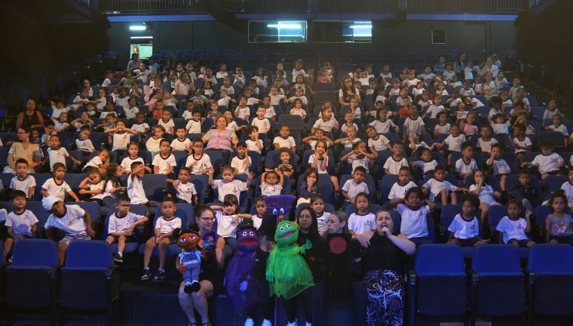 Fotografia mostra muitos meninos e meninas com uniforme da Prefeitura de SP sentados em um auditório estilo estádio. Na frente deles algunmsn adultos segurandos fantoches, sendo um deles um boneco ver e outro um semáforo com a luz vermelha evidente.