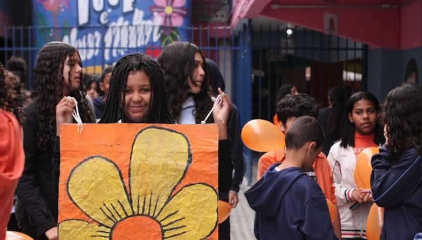 Fotografia de estudantes com balões laranja e cartaz de uma flor amarela com fundo roxo.