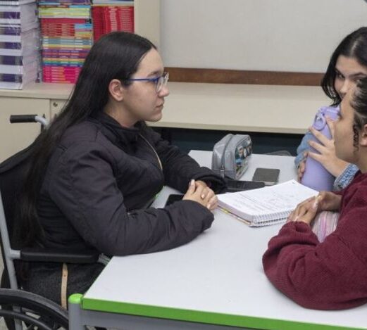 Foto de 3 meninas adolescentes em volta de uma mesa com livros ao fundo. Uma delas está na cadeira de rodas