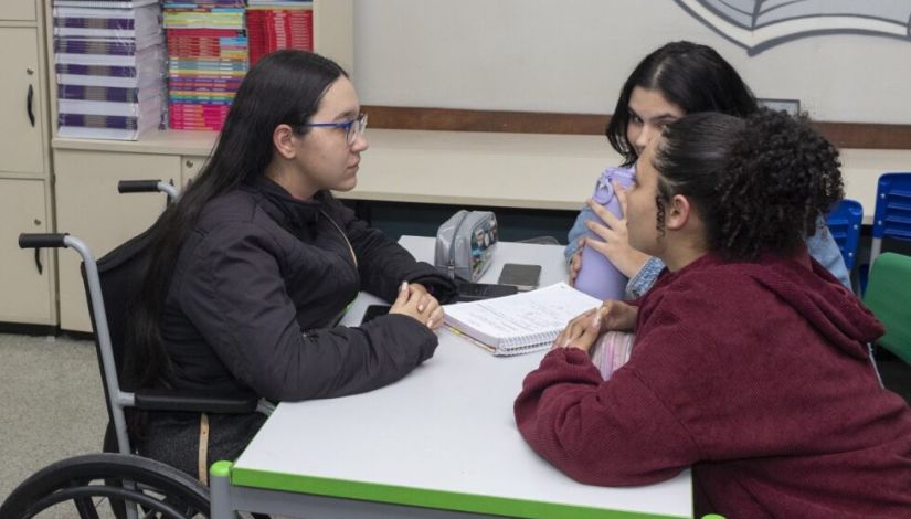 Foto de 3 meninas adolescentes em volta de uma mesa com livros ao fundo. Uma delas está na cadeira de rodas