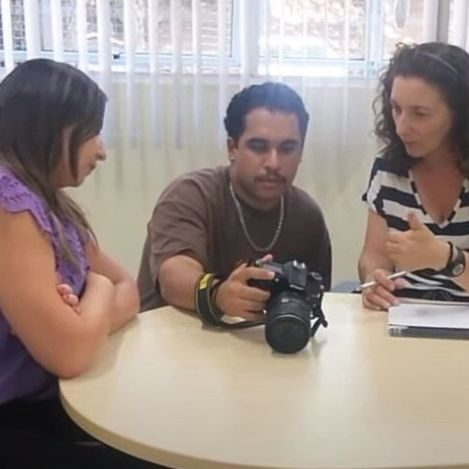 Foto de três pessoas, o homem, ao centro, segura uma máquina fotográfica e mostra as fotos para as duas mulheres