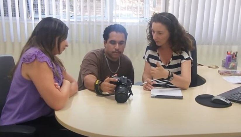 Foto de três pessoas, o homem, ao centro, segura uma máquina fotográfica e mostra as fotos para as duas mulheres