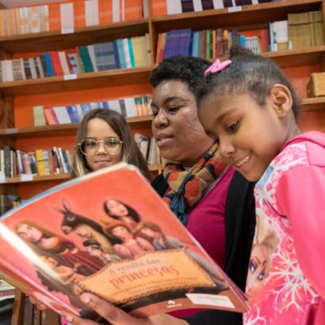 Foto de uma mulher lendo um livro para duas meninas. Ao fundo, prateleiras com livros