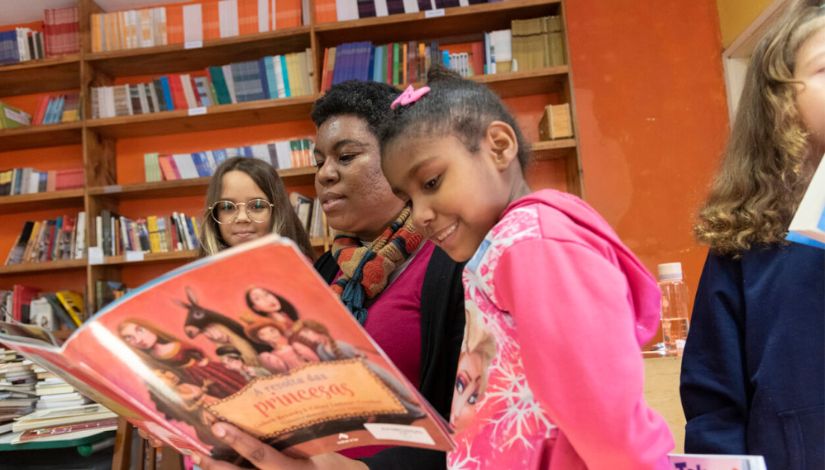 Foto de uma mulher lendo um livro para duas meninas. Ao fundo, prateleiras com livros