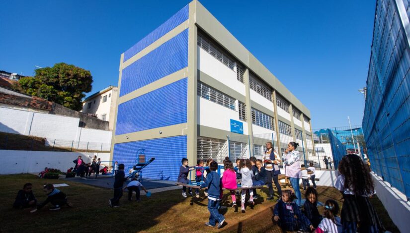 Imagem com um prédio de uma escola, na frente um gira gira com um grupo de crianças brincando