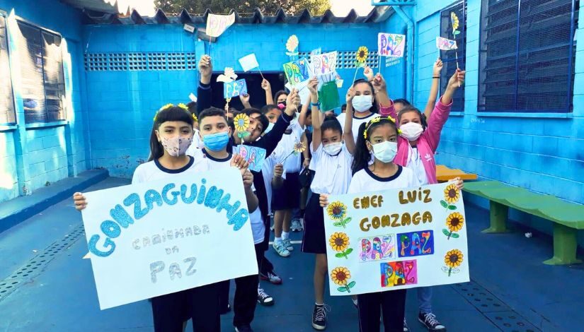 Fotografia de estudantes com cartazes nas mãos, bandeirinhas com a palavra paz. Alguns usam máscaras de proteção individual.