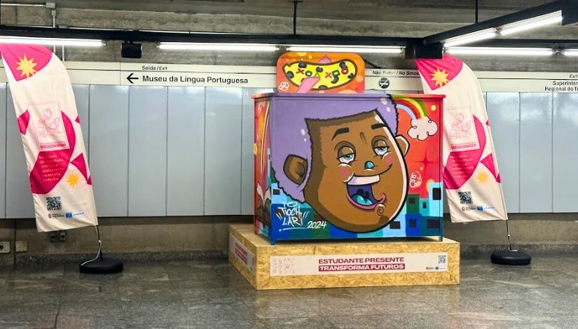 Fotografia de uma carteira e a cadeira em tamanho gigante que faz parte da campanha "Estudante Presente Transforma Futuros". A carteira está em uma estação de metrô. 