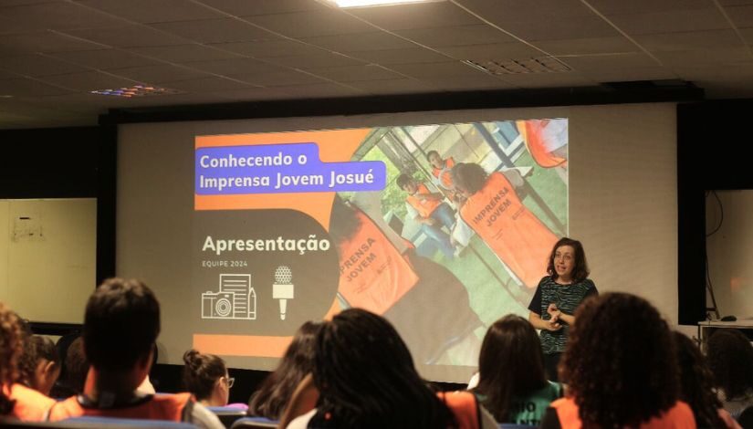 Fotografia de estudantes do Imprensa Jovem sentados em um auditório de costas para a fotografia e vendo uma professora a frente com uma tela projetada onde se lê 