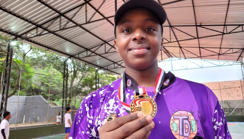 Estudante negro sorrindo, segurando sua medalha de ouro. Ele usa camiseta roxa e boné. 