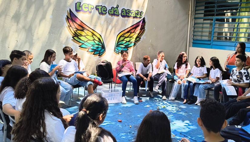 Fotografia de estudantes sentados em roda em momento de leitura. Ao fundo, na parede há uma pintura de par de asas onde se lê "Ler te dá asas". 