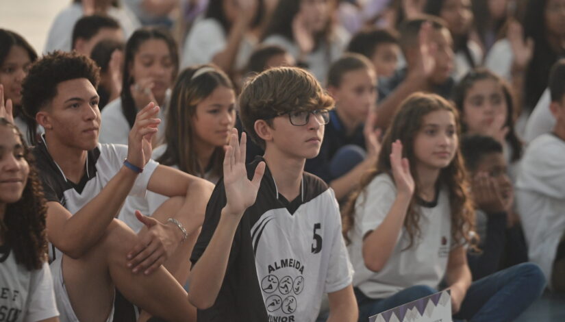 Muitos estudantes sentados no chão com a mão direita levantada.