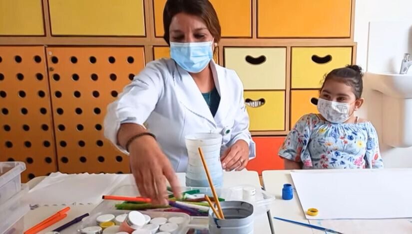 Fotografia de uma criança e uma professora elas fazendo atividade com folha branca, pincéis, tintas e canetinhas. Elas usam máscara de proteção individual.