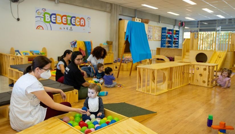 Fotografia mostra espaço de bebeteca, com brinquedos de madeira e bebês no chão explorando o espaço. Há mulheres adultas sentadas no chão auxiliando os bebês.