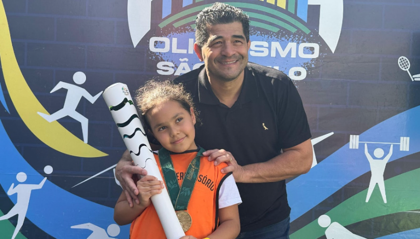 Um homem sorri para foto ao lado de uma criança com colete laranja segurando uma tocha olímpica e uma medalha