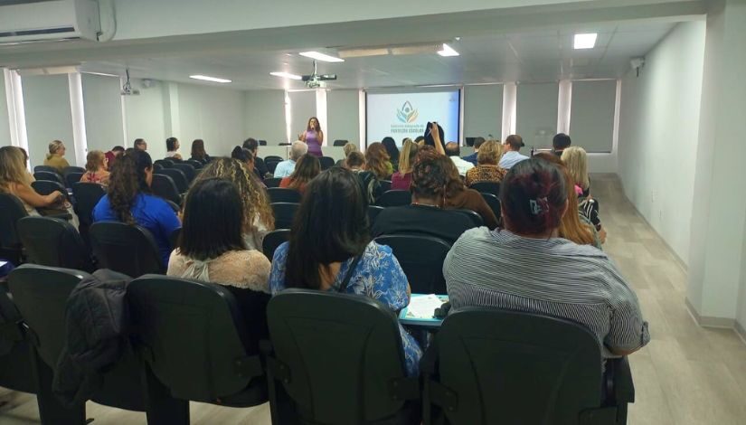 Fotografia mostra grupo de pessoas sentadas em auditório olhando para frente onde há uma mulher de pé segurando um microfone. Atrás dela há uma tela onde se lê "Gabinete Integrado de Proteção Escolar".