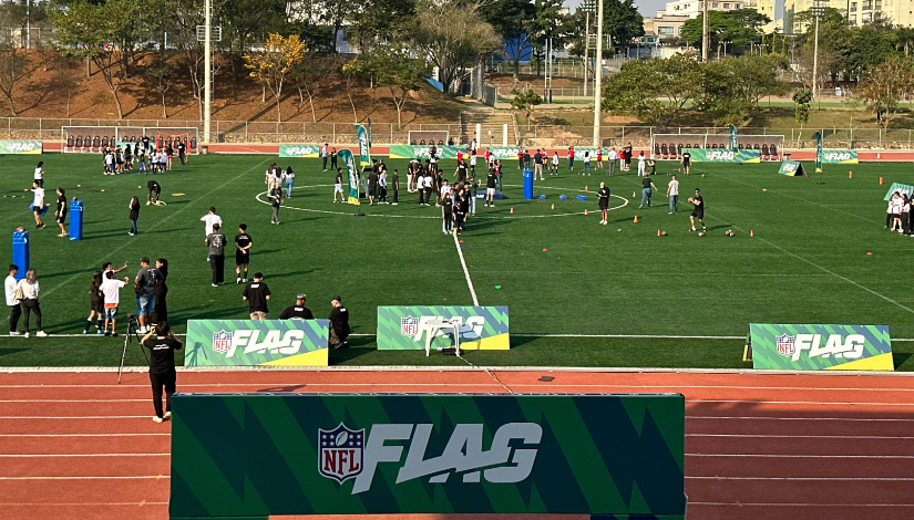 Imagem de um campo com pessoas praticanto futebol americano. Na frente um banner escrito 