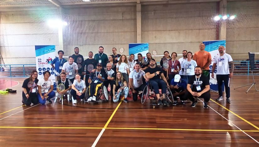 Grupo de pessoas estão posando para foto em um ginásio. Elas seguram raquetes de badminton.