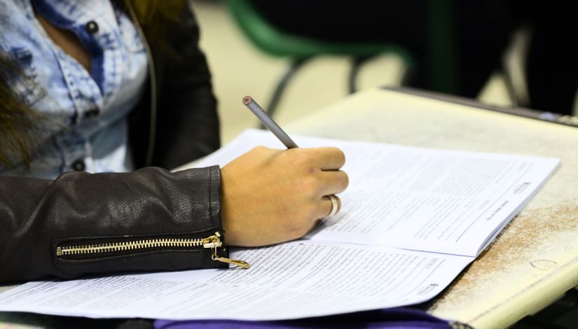 Imagem da mão de uma estudante segurando um lápis sobre um caderno de provas.