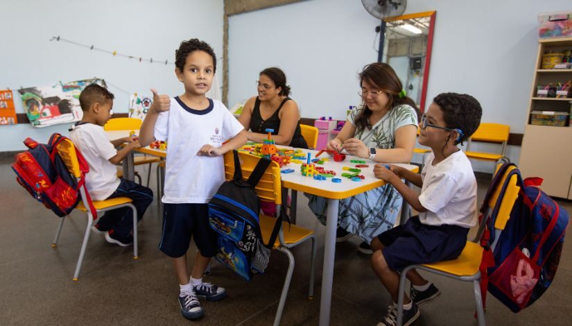 Imagem mostra estudantes sentados em volta de mesa e duas professoras sentadas com eles. Há um estudantes de pé olhando para a câmera.