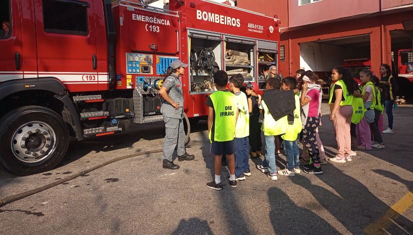 Fotografia mostra grupo de crianças usando colete verde com a escrita "DRE Ipiranga" de frente para um caminhão de bombeiros. Há uma bombeira mulher de pé na frente do caminhão conversando com as crianças.