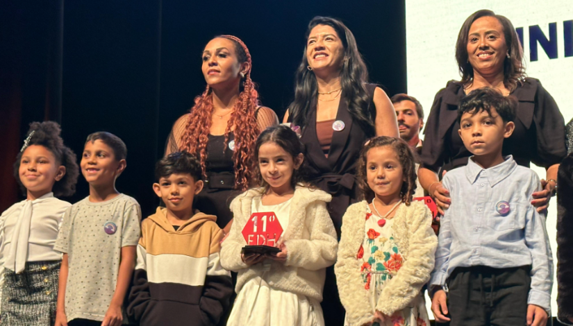 Imagem com um grupo de crianças e adultos em um palco segurando um trofeu vermelho escrito 