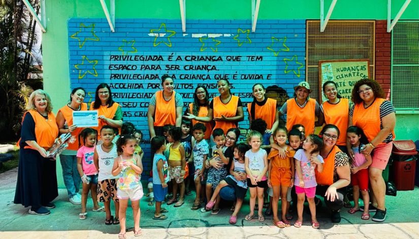 Crianças e adultos posam para foto, eles usam um colete laranja.