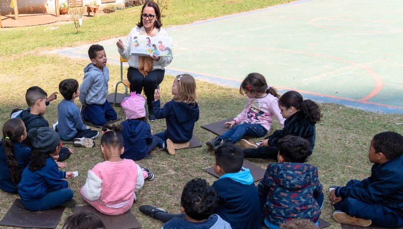 Fotografia de uma professora sentada em uma cadeirinha segurando um livro e a sua frente crianças sentadas em tapetinhos. Eles estão em uma área externa gramada com uma quadra ao fundo.