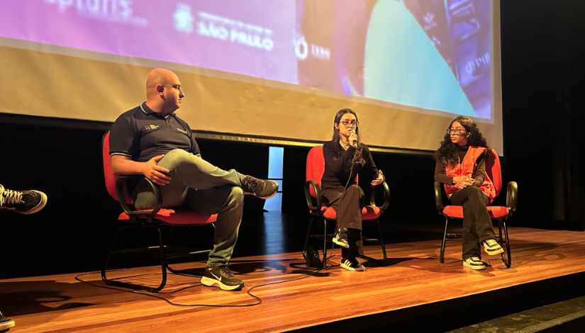 Três pessoas sentadas em cadeiras em um palco. Duas adolescentes, uma com roupas pretas e outra com um colete laranja. Ao lado, um homem com camiseta azul.