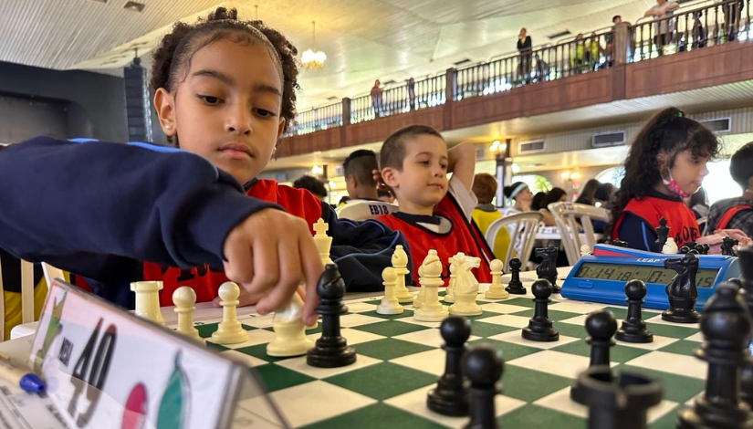 Uma imagem com uma estudante com uniforme da Prefeitura de São Paulo e um colete vermelho segurando um tabuleiro de Xadrez